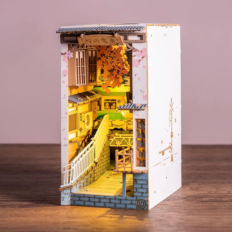 Op de afbeelding: Een miniatuur houten diorama van een Japanse straatsc&egrave;ne, verlicht met warme verlichting. De sc&egrave;ne omvat een brug, gebouwen en kersenbloesem accenten. Het diorama heeft de Japanse tekst "桜の電車" op een bord.