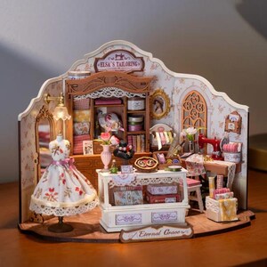 May include: A detailed miniature tailor's shop diorama. It features a dress form with a floral dress, a sewing machine, and shelves stocked with fabrics and haberdashery. The signs read "Elsa's Tailoring" and "Eternal Grace".
