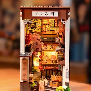 May include: A miniature Japanese street scene diorama with a detailed facade, featuring a shop with a sign in Japanese. The scene includes a lantern, cherry blossom accents, and various miniature items, creating a warm, inviting atmosphere.