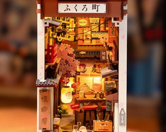 May include: A detailed miniature Japanese street scene diorama. The model features a shop with a Japanese sign, a lantern, and cherry blossom decorations. The scene is filled with miniature items, creating a warm and inviting atmosphere.