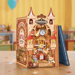 Op de afbeelding: Een houten boekenhoek met een miniatuur pretparkscène met een draaimolen, cadeauverpakkingen en een teddybeer. De boekenhoek heeft de tekst "Fairytale Amusement Park" en "Fun Never Closes". Een blauw boek staat ernaast.