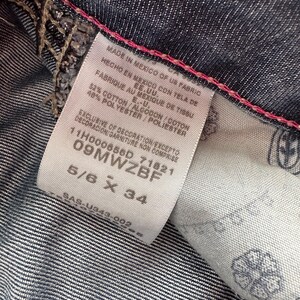 May include: A white tag with black text detailing the fabric content and origin of the jeans. The tag also includes the size 5/6 x 34. The tag is attached to a pair of dark blue denim jeans.