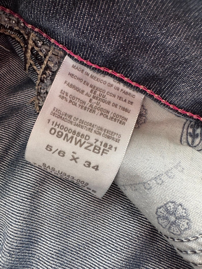 May include: A white tag with black text detailing the fabric content and origin of the jeans. The tag also includes the size 5/6 x 34. The tag is attached to a pair of dark blue denim jeans.