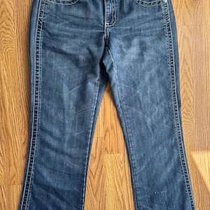 May include: A pair of dark blue denim jeans with a bootcut leg opening. The jeans have a light pink stitching detail along the seams and a small silver metal tag with the word "Wrangler" on the back waistband.