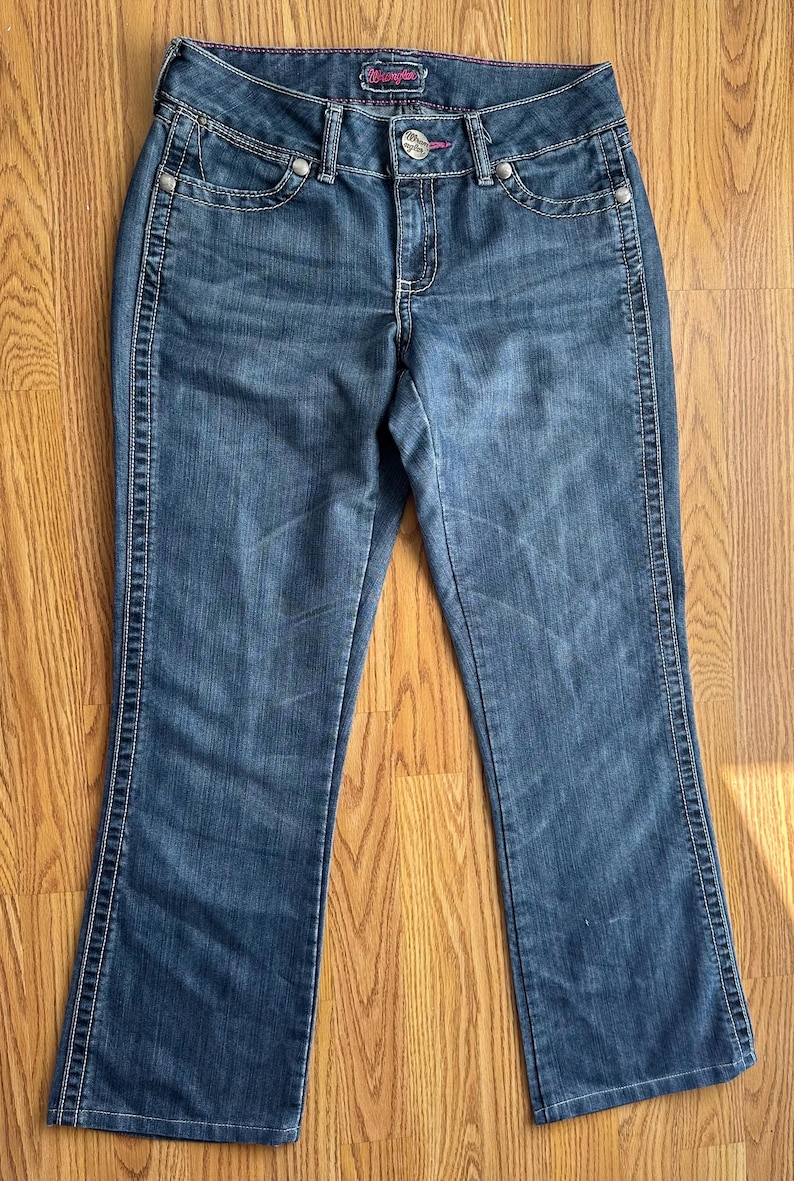 May include: A pair of dark blue denim jeans with a bootcut leg opening. The jeans have a light pink stitching detail along the seams and a small silver metal tag with the word "Wrangler" on the back waistband.