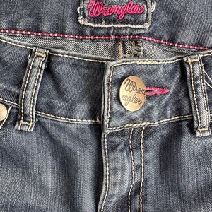 May include: Close-up of a pair of dark blue denim jeans with a Wrangler brand label and a metal button with the Wrangler logo.
