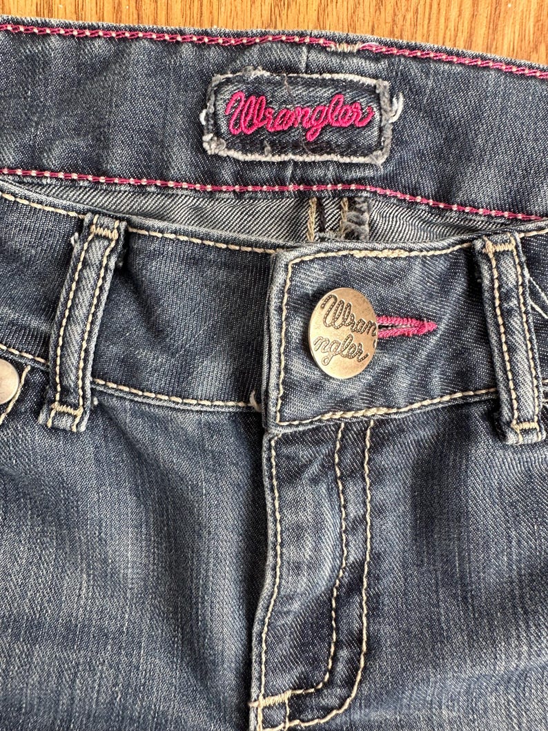 May include: Close-up of a pair of dark blue denim jeans with a Wrangler brand label and a metal button with the Wrangler logo.