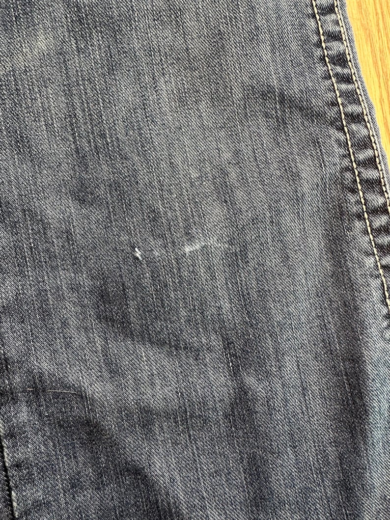 May include: Close-up of dark blue denim fabric with a subtle diagonal weave pattern. The fabric has a slightly worn look with a few faint white marks.