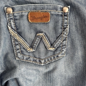 May include: A close-up of a blue denim pocket with a brown leather Wrangler label. The pocket has a decorative stitching design.
