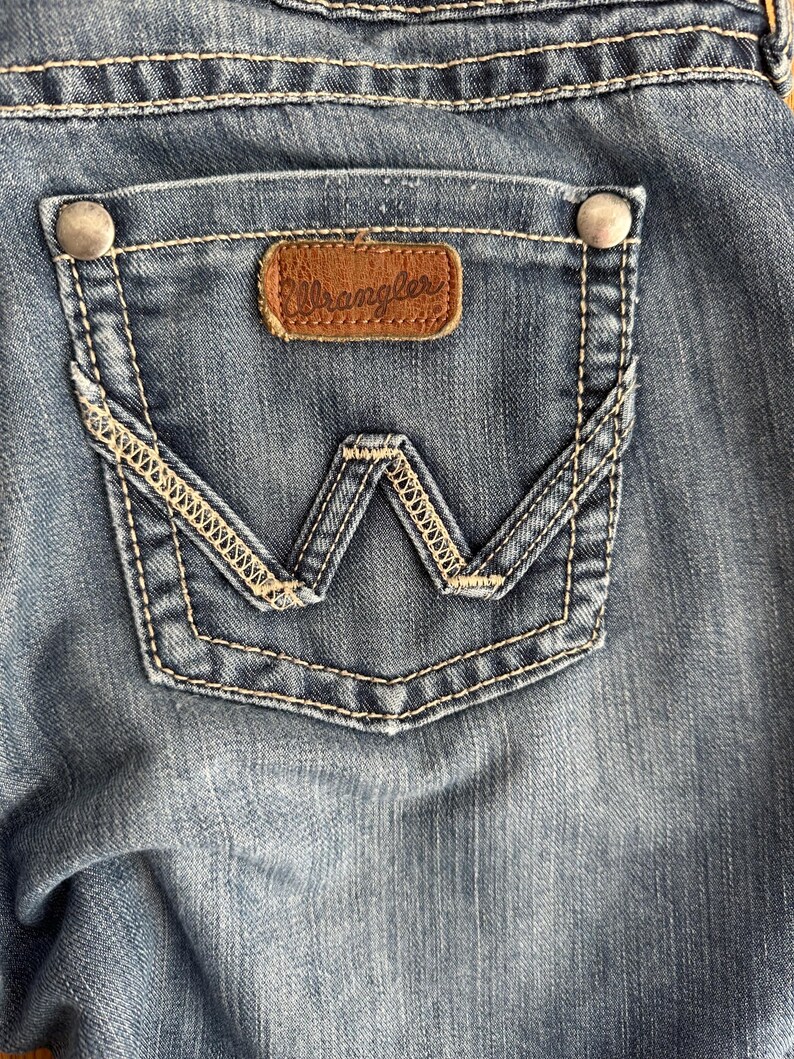 May include: A close-up of a blue denim pocket with a brown leather Wrangler label. The pocket has a decorative stitching design.
