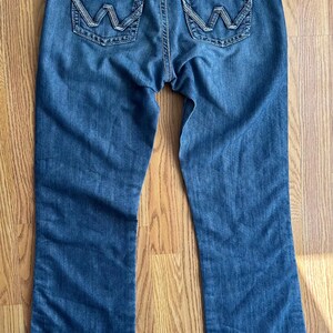 May include: A pair of blue denim jeans with a bootcut leg opening. The jeans have a light wash and feature a brown leather patch with the Wrangler logo on the back right pocket.