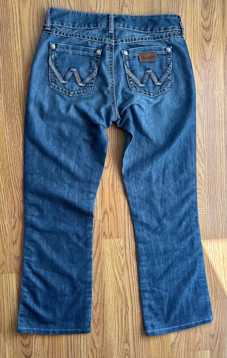 May include: A pair of blue denim jeans with a bootcut leg opening. The jeans have a light wash and feature a brown leather patch with the Wrangler logo on the back right pocket.