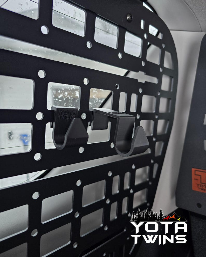 May include: Black metal cargo organizer with two black plastic hooks attached. The organizer has a grid pattern and is mounted in a vehicle. The text "YOTA TWINS" is visible in the lower right corner.