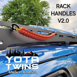 May include: Black roof rack with orange and yellow paracord handles. The text "RACK HANDLES V2.0" is displayed. The image also includes the "YOTA TWINS" logo and the text "Made in America". The rack is mounted on a blue vehicle.