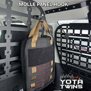 May include: A black molle panel hook with a tan strap is attached to a black metal cargo rack in a vehicle. A black bag with red and tan accents is hanging from the hook.