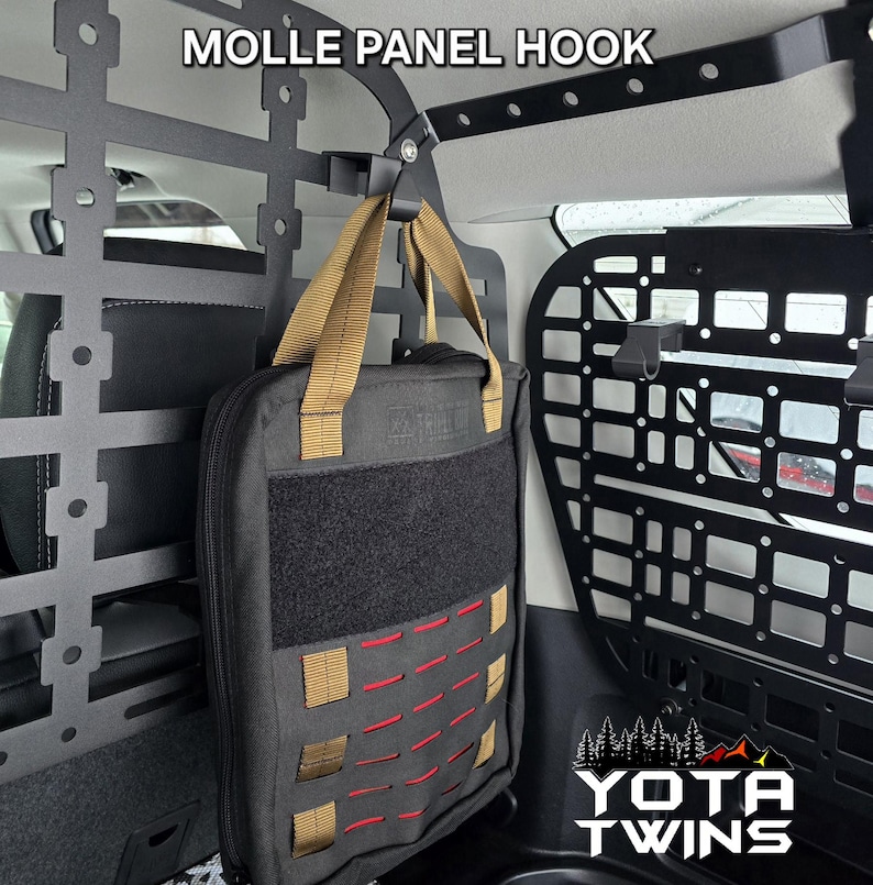 May include: A black molle panel hook with a tan strap is attached to a black metal cargo rack in a vehicle. A black bag with red and tan accents is hanging from the hook.