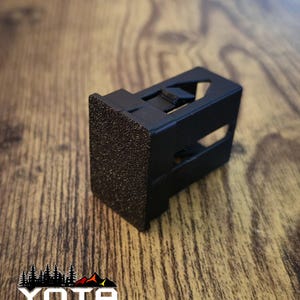 May include: A black plastic automotive trim clip with a textured surface. The clip has a small, rectangular shape and a locking mechanism.
