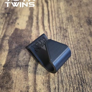 May include: A black plastic flush hook with the text "YOTA TWINS" embossed on the side. The hook is on a wooden surface.