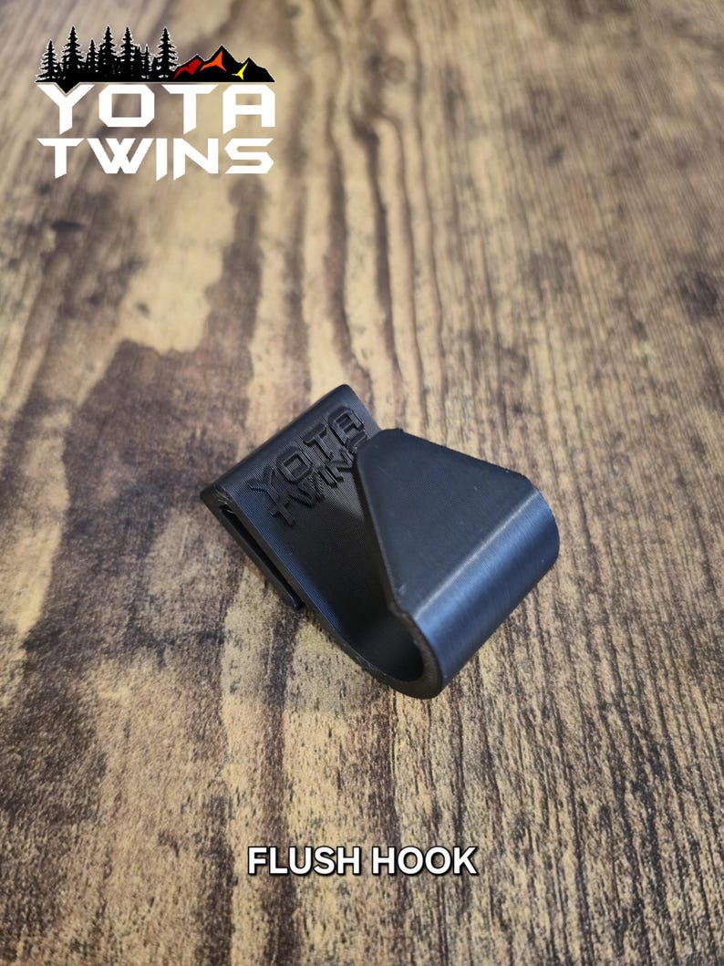 May include: A black plastic flush hook with the text "YOTA TWINS" embossed on the side. The hook is on a wooden surface.