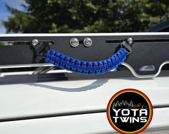 May include: A blue paracord grab handle attached to a black metal roof rack rail with silver bolts. The handle features a braided design and is mounted horizontally. A circular logo with the text YOTA TWINS is visible in the lower right corner.