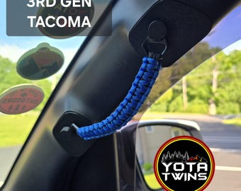 May include: A blue paracord grab handle installed on the A-pillar of a vehicle. The handle features a braided design and is attached to black mounting brackets. Text on the image reads 3RD GEN TACOMA and YOTA TWINS, with circular stickers visible.