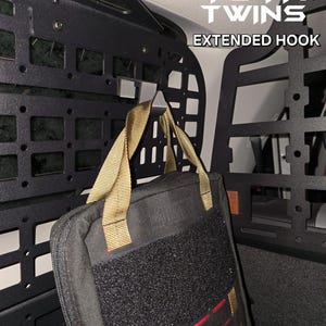 May include: A black metal extended hook mounted to a black metal cargo rack in a vehicle. A black bag with tan straps is hanging from the hook. The bag has a velcro patch on the front. The text "YOTA TWINS EXTENDED HOOK" is visible in the upper right corner of the image.