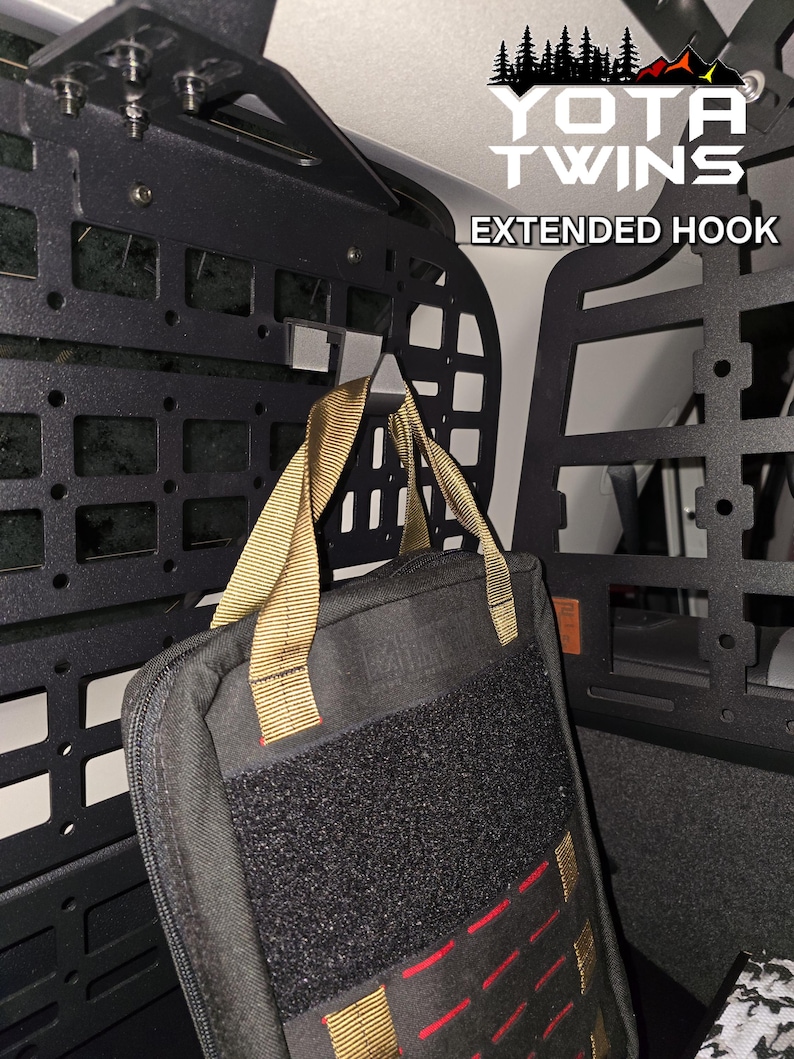 May include: A black metal extended hook mounted to a black metal cargo rack in a vehicle. A black bag with tan straps is hanging from the hook. The bag has a velcro patch on the front. The text "YOTA TWINS EXTENDED HOOK" is visible in the upper right corner of the image.