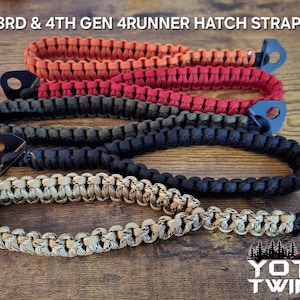 May include: Four different coloured paracord straps with black metal clasps. The straps are in the colours orange, red, black, and tan. The straps are labelled "3RD & 4TH GEN 4RUNNER HATCH STRAP" and "YOTA TWINS".