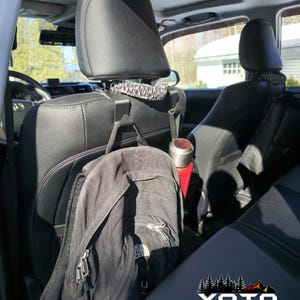 May include: A pair of black headrest seat hooks installed in the backseat of a car. A black backpack and a red water bottle are hanging from the hooks. The car seat is gray and black. The logo "YOTA TWINS" is visible on the seat.