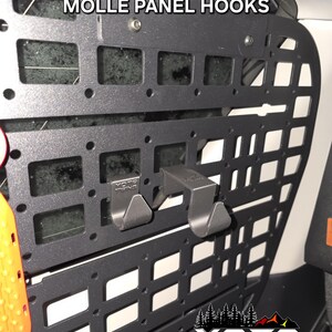 May include: Black metal MOLLE panel with two black hooks attached. The panel has a grid pattern and the text "MOLLE PANEL HOOKS" is printed on the top. The text "YOTA TWINS" is printed on the bottom of the panel.