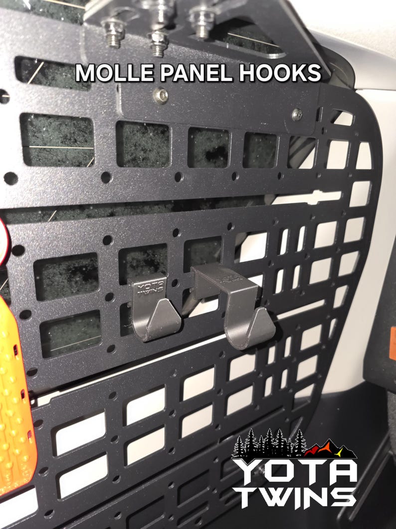 May include: Black metal MOLLE panel with two black hooks attached. The panel has a grid pattern and the text "MOLLE PANEL HOOKS" is printed on the top. The text "YOTA TWINS" is printed on the bottom of the panel.