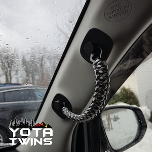 Peut inclure: Une poignée en paracorde noir et blanc est fixée à l'intérieur d'une voiture. La poignée est fixée à deux crochets en plastique noir. Le texte "SRS CURTAIN AIRBAG" est visible à l'intérieur de la voiture.