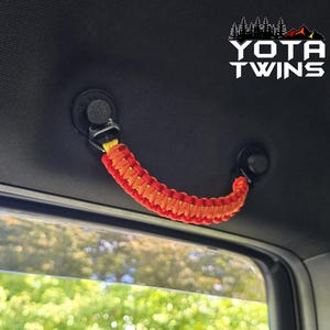 May include: A vehicle grab handle made of braided red and yellow paracord, attached to a black interior ceiling mount. The handle features a textured, woven design and is positioned above a window. The text YOTA TWINS is visible in the corner.