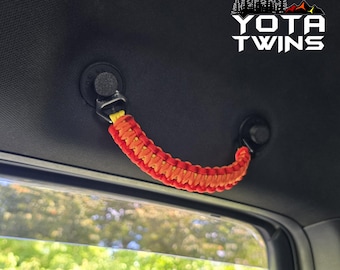 May include: A vehicle grab handle made of braided red and yellow paracord, attached to a black interior ceiling mount. The handle features a textured, woven design and is positioned above a window. The text YOTA TWINS is visible in the corner.