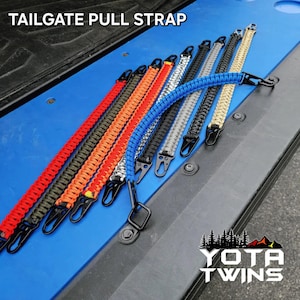 May include: A set of ten colorful paracord tailgate pull straps with black metal clips. The straps are made of braided paracord and are available in a variety of colors, including red, orange, green, blue, and black. The straps are labeled "TAILGATE PULL STRAP" and "YOTA TWINS".
