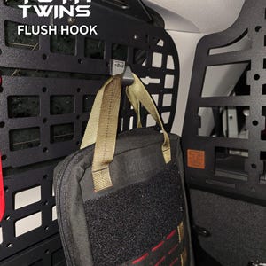 May include: A black metal flush hook with a black and tan bag hanging from it. The hook is attached to a black metal panel with the words "YOTA TWINS FLUSH HOOK" printed on it.
