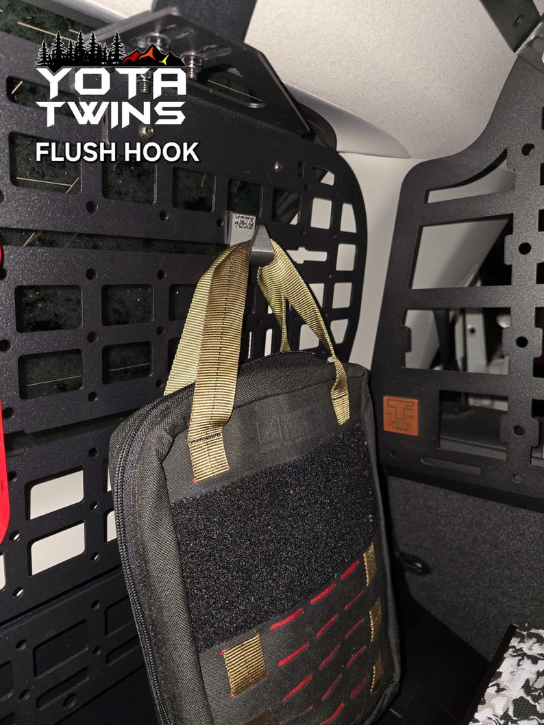 May include: A black metal flush hook with a black and tan bag hanging from it. The hook is attached to a black metal panel with the words "YOTA TWINS FLUSH HOOK" printed on it.