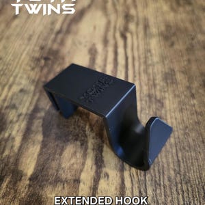 May include: Black plastic extended hook with the text "YOTA TWINS" printed on it. The hook is sitting on a wooden surface.