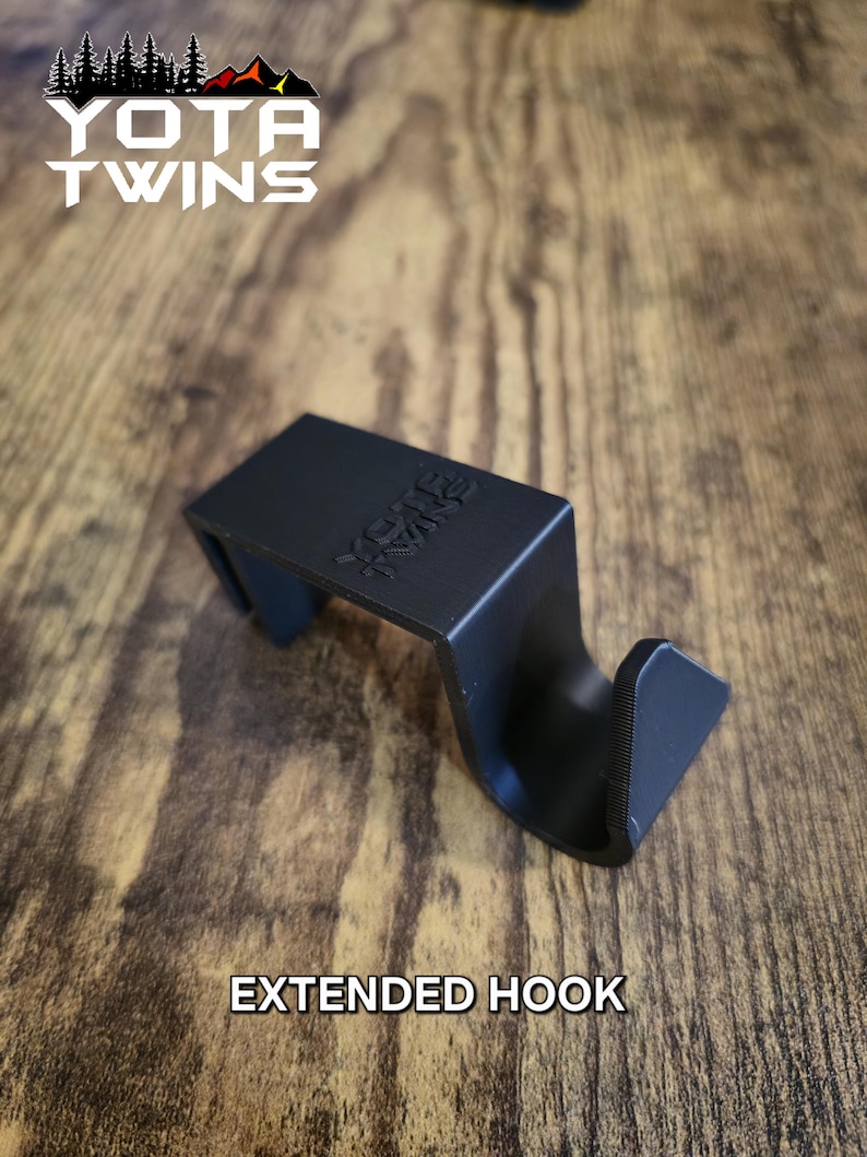 May include: Black plastic extended hook with the text "YOTA TWINS" printed on it. The hook is sitting on a wooden surface.