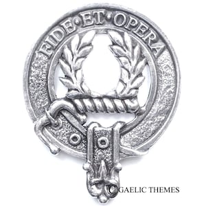 Op de afbeelding: Een zilverkleurig tinnen clanwapen met de tekst "Fide Et Opera" rond de bovenrand. Het wapen heeft een krans van bladeren, een touw en een band met twee cirkels. De onderkant van het wapen heeft een decoratief ontwerp.