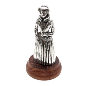 May include: A silver figurine of a woman wearing a ruff collar and a hat, holding a rosary. The figurine is standing on a wooden base.
