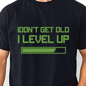 May include: A black short-sleeved t-shirt featuring green pixelated text that reads "I DON'T GET OLD I LEVEL UP" above a green loading bar graphic. The design is centered on the chest of the garment, which is worn with light blue denim jeans.