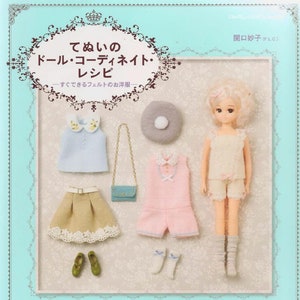 May include: A book cover with a light blue background and a white border. The title is "てぬいの ドール・コーディネイト・レシピ" which translates to "Handmade Doll Coordinate Recipe". The book features a doll with blonde hair wearing a white and pink outfit and a light blue and white outfit. The doll is holding a small white purse.