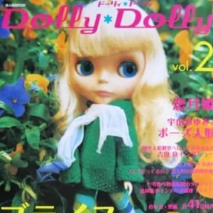 May include: A magazine cover featuring a doll with blonde hair and blue eyes wearing a green sweater and a white scarf. The magazine is titled "Dolly*Dolly" and the cover text reads "vol. 2" and "ブライスと新世代ファッションドールたち".