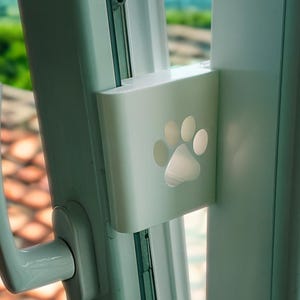 Op de afbeelding: Een witte raamvergrendeling met een uitsparing in de vorm van een pootafdruk, geïnstalleerd op een wit raamkozijn. Het raam staat gedeeltelijk open en biedt uitzicht op een groen landschap en een blauwe lucht. De raamkruk is zichtbaar.