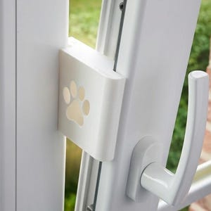 Peut inclure: Un verrou de fenêtre blanc avec un motif de patte de chien. Le verrou est monté sur un cadre de fenêtre blanc, avec une poignée visible à droite. L'arrière-plan montre un jardin vert et une maison.