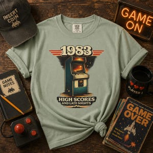 Könnte beinhalten: Ein salbeigrünes T-Shirt mit einer Grafik eines Vintage-Spielautomaten und dem Text 1983 sowie HIGH SCORES AND LATE NIGHTS. Das Design enthält rote horizontale Streifen und einen Sternenhintergrund, gedruckt auf einem kurzärmeligen Rundhals-T-Shirt.