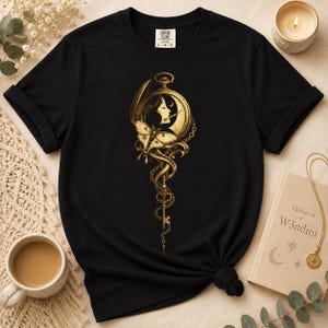 Może przedstawiać: Czarny t-shirt z krótkim rękawem z nadrukiem w kolorze złotym, przedstawiającym zegarek kieszonkowy z profilem osoby, ćmą oraz długim, ozdobnym kluczem na łańcuszku. Koszulka posiada metkę Comfort Colors przy wewnętrznym kołnierzyku.