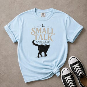 Puede incluir: Una camiseta de manga corta de color azul claro con un gráfico de un gato negro y el texto "SMALL TALK CANCELLED." El diseño incluye un pequeño icono de luna creciente sobre el texto. La camiseta tiene cuello redondo y corte estándar.