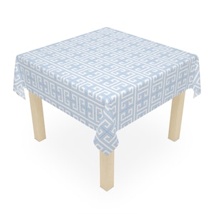 May include: A square tablecloth featuring a repeating white Greek key pattern on a light blue background, draped over a table with four light-colored wooden legs. The fabric covers the top and sides of the table, displaying a geometric design.
